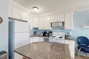 A kitchen with white cabinets, a granite countertop, a white stove, a microwave, and a blue chair. - Seascape Condos #1219 (Galveston)