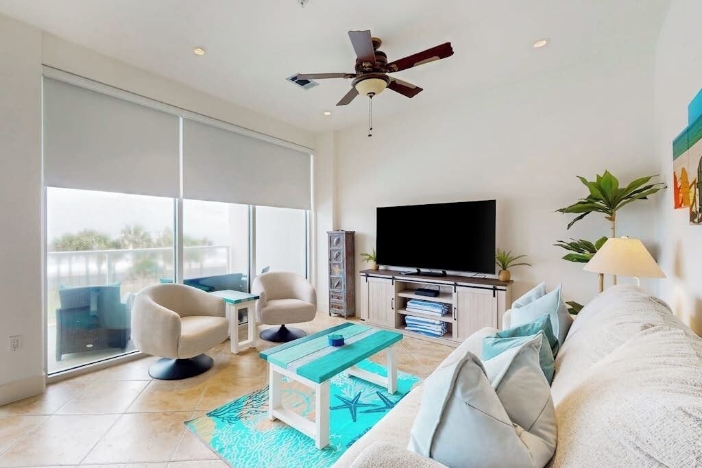 A modern living room with a flat-screen TV, a ceiling fan, a sofa, a coffee table, and a large window.