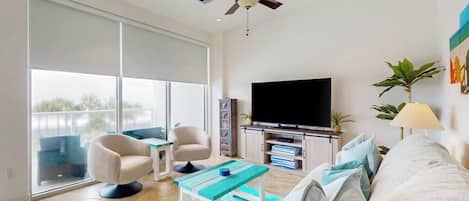 A modern living room with a flat-screen TV, a ceiling fan, a sofa, a coffee table, and a large window.