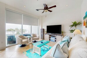 Un salon moderne doté d’un téléviseur à écran plat, d’un ventilateur de plafond, d’un canapé, d’une table basse et d’une grande fenêtre.