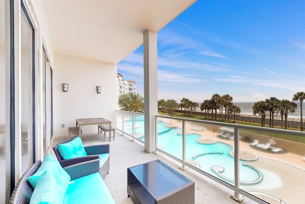 A balcony with wicker furniture, a glass table, and a view of a pool area with palm trees and a beach.