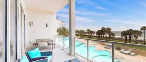 A balcony with wicker furniture, a glass table, and a view of a pool area with palm trees and a beach.