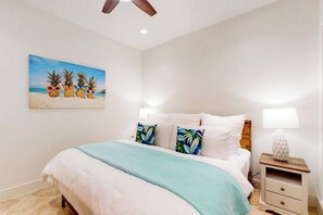 A bedroom with a bed, pillows, a nightstand, a lamp, and a painting of pineapples on the wall. - Pelican Oasis - Beachfront Condo, Pool & Hot Tub (Galveston)