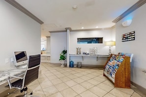 A reception area with a desk, computer, chair, plant, and a magazine rack. - Pelican Oasis - Beachfront Condo, Pool & Hot Tub (Galveston)