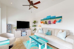 A living room with a flat-screen TV, a ceiling fan, a sofa, a coffee table, and a wall art piece. - Pelican Oasis - Beachfront Condo, Pool & Hot Tub (Galveston)