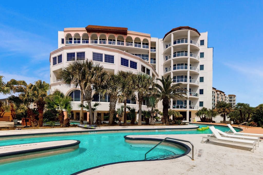 A multi-story hotel with a pool area, palm trees, and a clear blue sky.