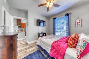 A bedroom with a bed, a ceiling fan, a window with blinds, a TV on the wall, and a kitchen area visible through an open door. - Beach Club 155 - Pool & Hot Tub, Close to Beach (Corpus Christi)
