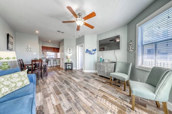 A spacious living area with a ceiling fan, a dining table with chairs, a kitchen in the background, and a large window with blinds. - Beach Club 155 - Pool & Hot Tub, Close to Beach (Corpus Christi)