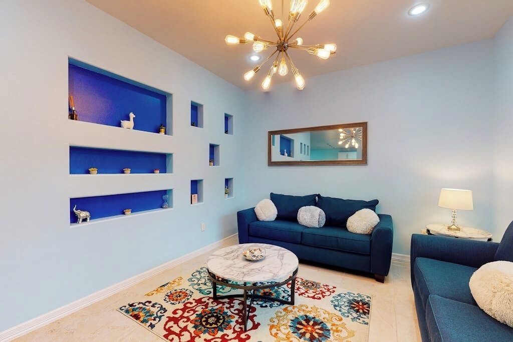A living room with a blue sofa, a round coffee table, and a patterned rug. There are shelves with decorative items and a ceiling light fixture.