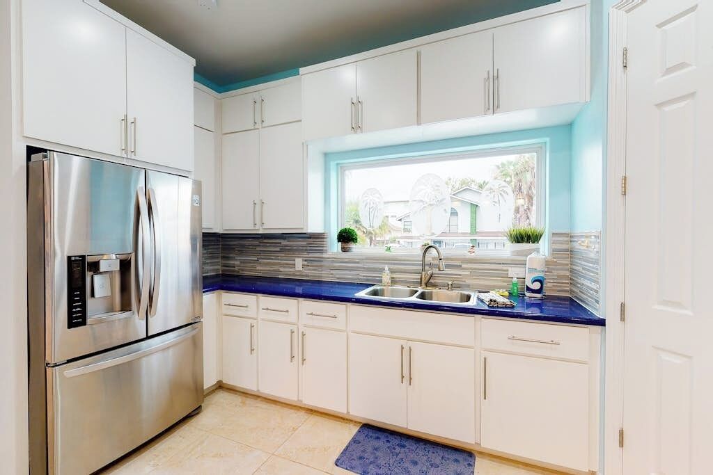 A modern kitchen with white cabinets, a stainless steel refrigerator, a blue countertop, and a window with a view of the outside.