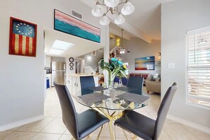 Uma sala de jantar moderna com mesa de vidro e cadeiras pretas, cozinha ao fundo e obras de arte nas paredes.