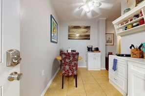 A kitchen with white cabinets, a dining table with a floral chair, and a ceiling fan. - White Granite at 106 East Huisache (South Padre Island)