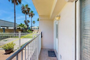 A balcony with a white railing, a door, and a view of palm trees and a building in the background. - Dolphin Condominiums #5 (South Padre Island)