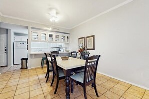 A dining area with a table and chairs, a trash can, and a kitchen area with cabinets and appliances. - Dolphin Condominiums #5 (South Padre Island)