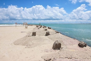 A beach with a rocky breakwater extending into the sea, a signboard, and a person standing at the end of the breakwater. - Dolphin Condominiums #5 (South Padre Island)