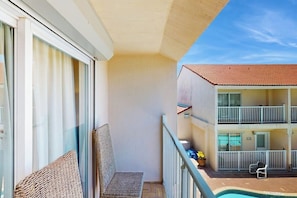 A balcony with a wicker chair, a poolside area with a chair and a table, and a building with a red-tiled roof. - Dolphin Condominiums #5 (South Padre Island)
