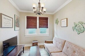 Un salon comprenant un canapé, un banc, une table basse, un téléviseur et un lustre. Il y a deux fenêtres équipées de stores, et on peut voir des éléments de décoration murale.