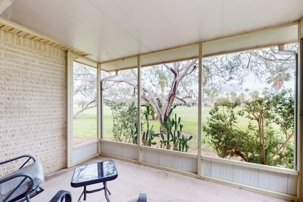 Une terrasse couverte avec un mur en briques, une porte vitrée et une vue sur un espace verdoyant avec des arbres.