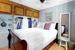 A bedroom with a wooden bed frame, a white bedspread, and decorative pillows. There is a window with blue curtains, a painting on the wall, and a small table with a mirror. - Long Island Village 790 W Oyster Drive Bungalow (Port Isabel)