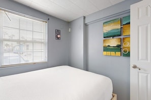 A bedroom with a bed, a window with blinds, and a wall with artwork. - Long Island Village 790 W Oyster Drive Bungalow (Port Isabel)