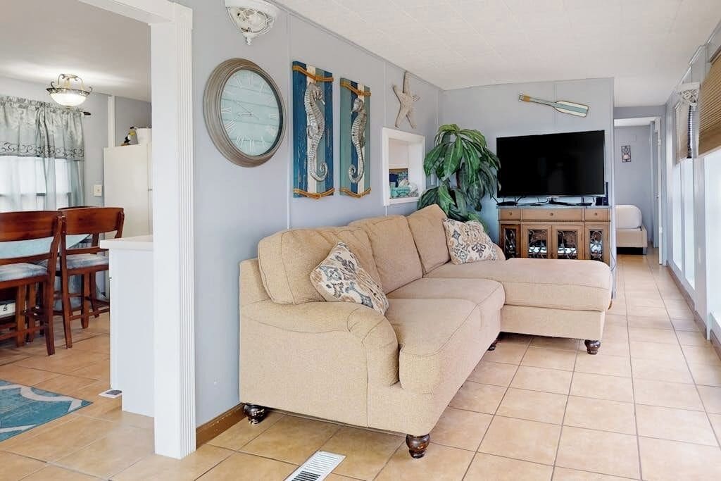 Una sala de estar con un sofá seccional color beige, un comedor con sillas de madera, un televisor y decoración en la pared.