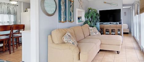 Una sala de estar con un sofá seccional color beige, un comedor con sillas de madera, un televisor y decoración en la pared.