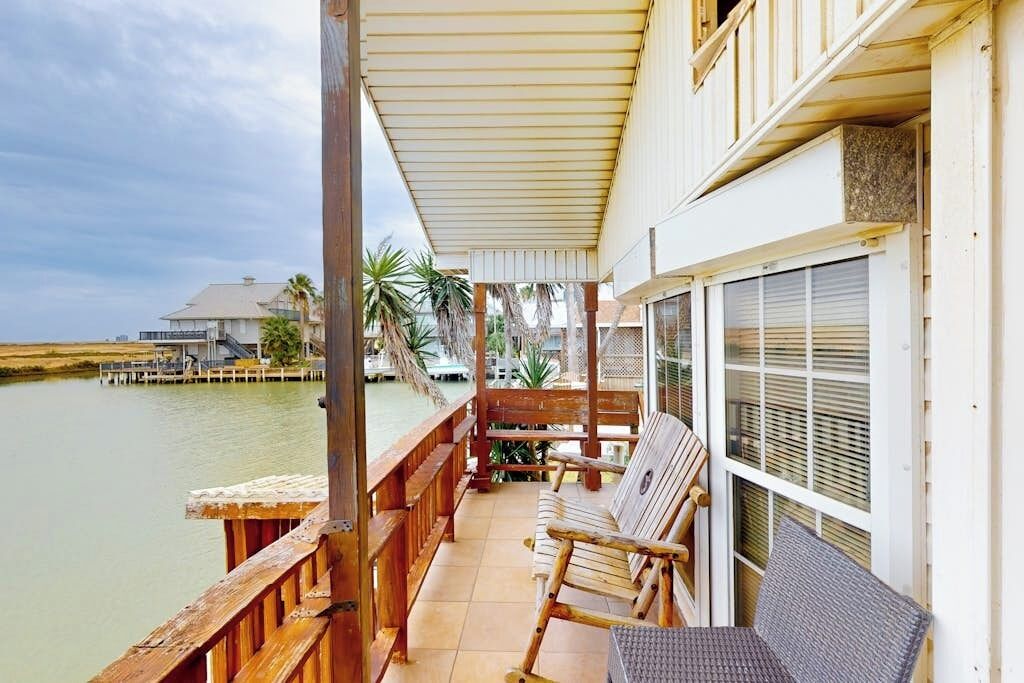 A wooden deck with a covered seating area overlooking a body of water, a building with a large window, and a palm tree in the background.