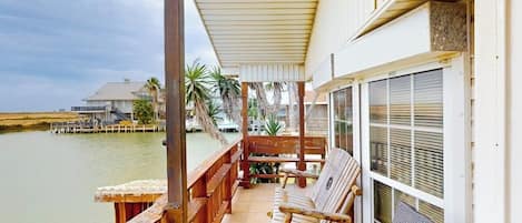 A wooden deck with a covered seating area overlooking a body of water, a building with a large window, and a palm tree in the background.