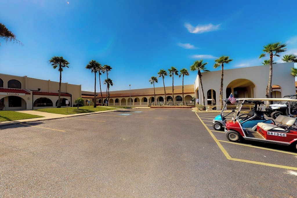 Un parking avec un véhicule de type voiturette de golf, des palmiers et un bâtiment doté d’arcades.