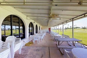An outdoor covered patio with white plastic chairs and tables, large windows, and a ceiling fan. - Long Island Village 790 W Oyster Drive Bungalow (Port Isabel)