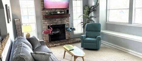 Een woonkamer met een bank, een smaragdgroene fauteuil, een salontafel, een open haard, een televisie en een groot raam.
