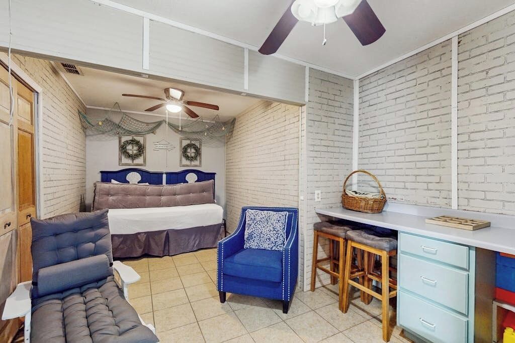 Ein Zimmer mit einem Bett, einem blauen Sessel, einem Schreibtisch mit Hocker und einem Deckenventilator.