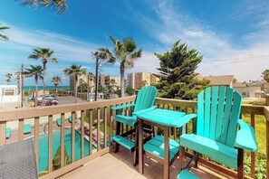 Ein Balkon mit türkisfarbenen Adirondack-Stühlen und einem Holztisch mit Blick auf einen Swimmingpool und eine Straße mit Palmen und Gebäuden.