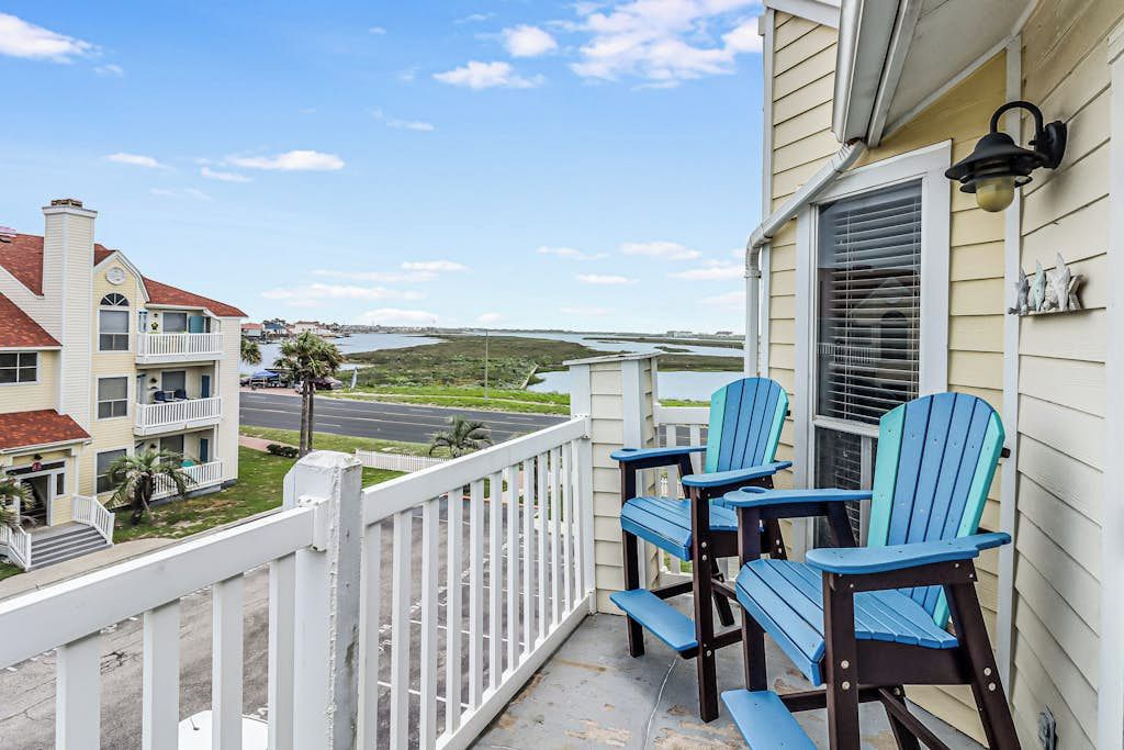 Un balcon avec deux fauteuils Adirondack, une balustrade blanche et une vue sur une étendue d’eau et des bâtiments.