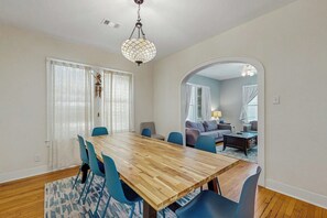 A dining area with a wooden table and blue chairs, a chandelier, and a view into a living room through an archway. - Sea Glass Cottage - Dog-Friendly, Near the Beach (Galveston)