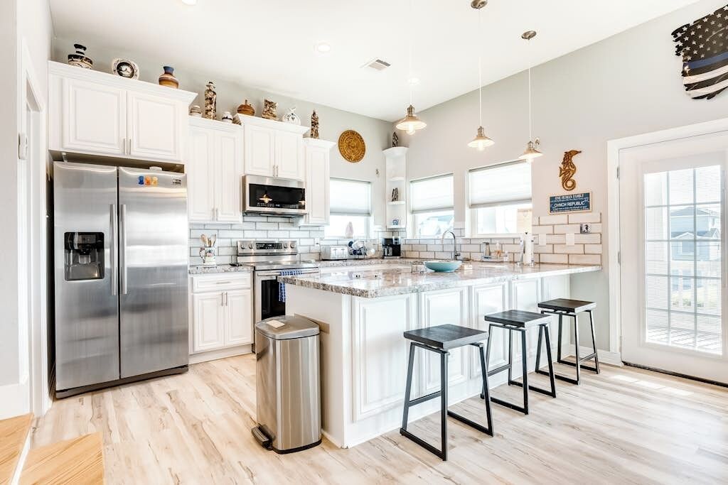 A modern kitchen with white cabinetry, stainless steel appliances, a large island with bar stools, and a view of the city through the windows.