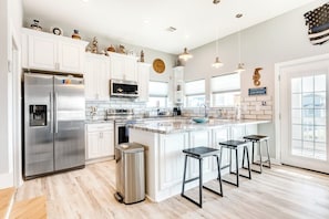 A modern kitchen with white cabinetry, stainless steel appliances, a large island with bar stools, and a view of the city through the windows. - Seahorse Beach House (Galveston)