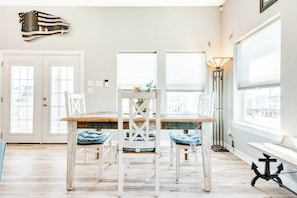 A dining area with a wooden table and white chairs, a large window with blinds, and a floor lamp. - Seahorse Beach House (Galveston)
