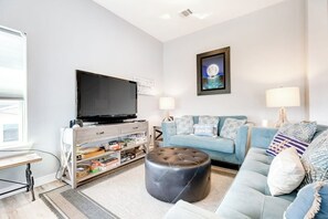 A modern living room with a flat-screen TV, a light blue sofa, a round ottoman, and a framed picture on the wall. - Seahorse Beach House (Galveston)