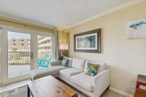 A living room with a sofa, a coffee table, and a view of a balcony with a chair. - Victorian Condos Barefoot Beach Haven (Galveston)