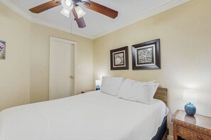 Ein Hotelzimmer mit Bett, Deckenventilator, Nachttisch mit Lampe und zwei gerahmten Bildern an der Wand.