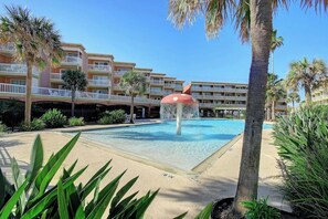 A hotel pool area with a water feature, palm trees, and a multi-story building in the background. - Victorian Condos Barefoot Beach Haven (Galveston)