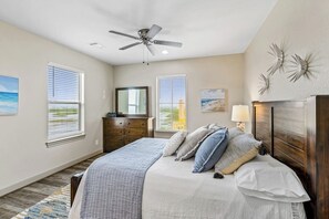 A bedroom with a bed, a ceiling fan, a dresser, a mirror, and artwork on the walls. - Pointe West - Bay Breeze Paradise (Galveston)