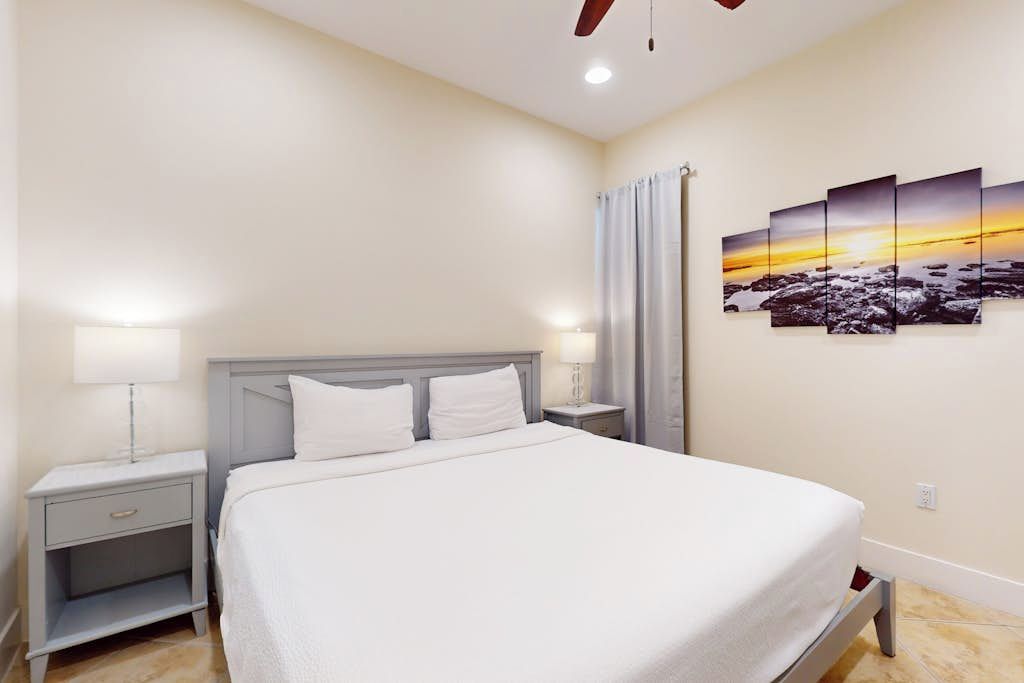 A hotel room with a bed, nightstands, a lamp, a ceiling fan, and a sunset painting on the wall.