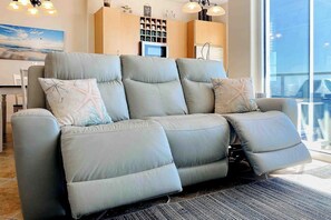 Ein graues, leicht zurückgelehntes Sofa mit vielen Kissen, ein großes Fenster mit Ausblick und eine Küchenzeile im Hintergrund.
