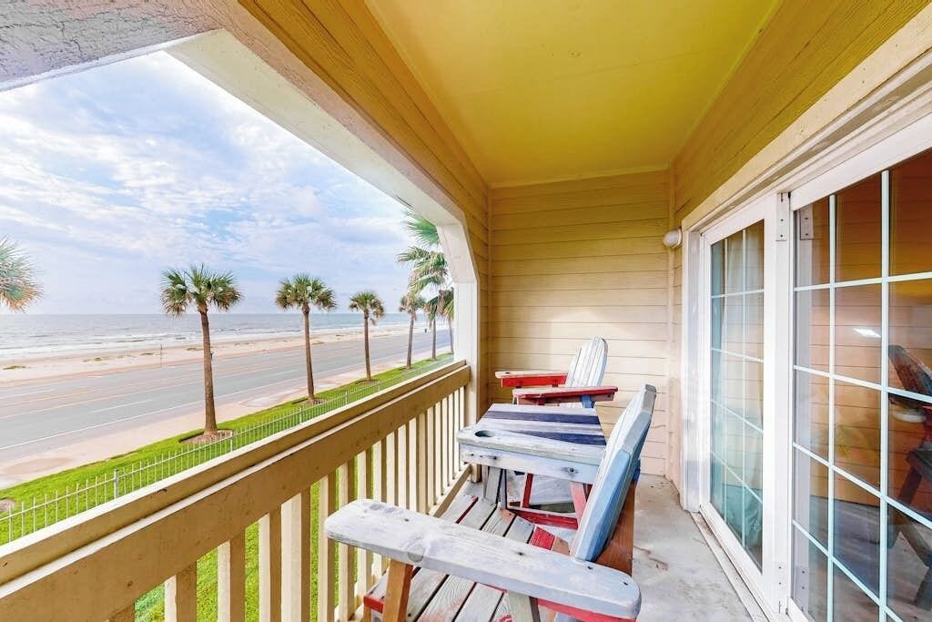 A balcony with a view of the beach, wooden railing, and two Adirondack chairs.