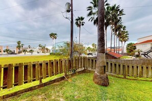 A fenced yard with a palm tree, a wooden fence, and a building in the background. - Aztec Habitat No 19 - Pool, 1 Minute to the Beach (South Padre Island)