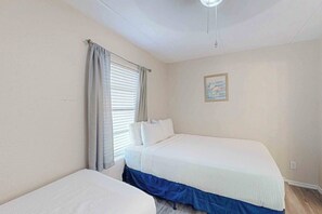 Ein Schlafzimmer mit zwei Betten, einem fensterbelaubten Fenster, einem Deckenventilator und einem gerahmten Bild an der Wand.