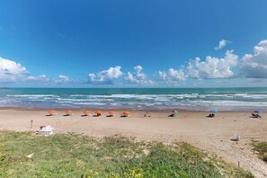A beach with umbrellas and people, a clear sky, and the ocean. - Inverness #304 - Steps to Beach, Pool & Hot Tub (South Padre Island)