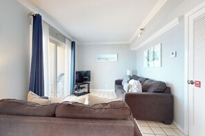 A living room with a sofa, a television, and a window with curtains. - Inverness #304 - Steps to Beach, Pool & Hot Tub (South Padre Island)